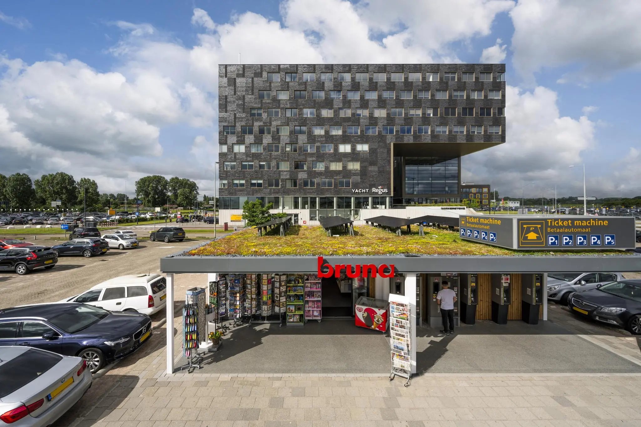 Modern kantoorgebouw bij Rotterdam The Hague Airport met op de voorgrond een Bruna-winkel en parkeerautomaten.
